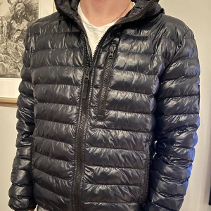 Svart pufferjacka med huva - Snygg svart pufferjacka med huva och dragkedja framtill. Jackan har en quiltad design och en praktisk bröstficka med dragkedja. Perfekt för kyligare dagar.