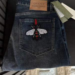 Mörkblåa jeans från Gucci  - Snygga mörkblå jeans från Gucci med en unik broderad insekt på bakfickan. Jeansen har en klassisk femficksdesign och en knappgylf. Perfekta för dig som vill ha något extra i garderoben.