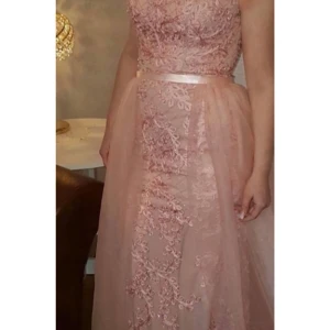 Rosa spetsklänning - Elegant rosa långklänning med spetsdetaljer och ett satinband i midjan. Klänningen har en vacker broderad design och är perfekt för festliga tillfällen.