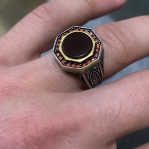 Unik ring med röd sten och detaljer - Säljer en äkta silver unik ring med en stor röd sten i mitten, omgiven av små orange stenar. Ringen har detaljerade mönster på sidorna och är tillverkad i silver med en guldliknande kant runt stenen. Perfekt för att ge en touch av stil och elegans.