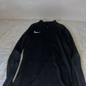 Svart långärmad tröja från Nike - Snygg svart långärmad tröja från Nike med dragkedja vid halsen och mönstrade ärmar. Perfekt för träning eller en avslappnad dag. Den har en stilren design med Nike-loggan på bröstet.