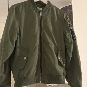 Bomberjacka  - Säljer en snygg grön bomberjacka från H&M med dragkedja framtill och en cool patch på ärmen. Jackan har ribbade muddar och en ficka med dragkedja på ärmen. Perfekt för en casual look! Jag skulle säga att den är XS/S