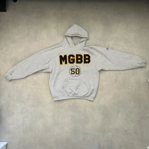 Grå hoodie med tryck - Säljer en grå hoodie med tryck 'MGBB 50' i svart och gult på framsidan. Den har en broderad text 'SVENSSON' på huvan och '2003' på ärmen. Perfekt för en avslappnad stil. Från märket LEE 