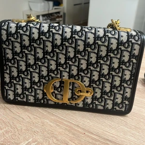 Mönstrad axelväska med gulddetaljer - Snygg axelväska med monogrammönster och guldfärgad logodetalj framtill. Väskan har en matchande liten plånbok och en dragkedja inuti. Perfekt för att hålla dina saker organiserade och stiliga. Kan säljas utan plånbok om så önskas