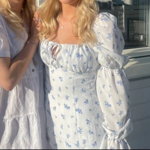 Vit blommig klänning  - Säljer denna superfina klänning från Divided H&M i storlek 36. Klänningen är vit med små blåa blommor, puffärmar med sömmar samt att den har en dragkedja vid sidan. 