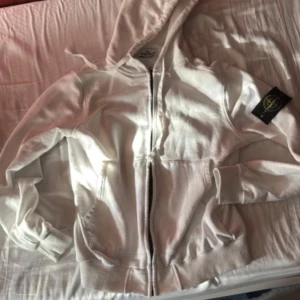 Vit zip up hoodie från Stone Island - Säljer en stilren vit huvtröja från Stone Island med dragkedja och den ikoniska loggan på ärmen. Perfekt för en casual look. Tröjan har långa ärmar och en bekväm passform.