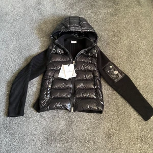 Svart dunjacka från Moncler - Snygg svart dunjacka från Moncler med huva och dragkedja. Jackan har stickade ärmar och en quiltad kropp med en ficka på ärmen. Perfekt för kyligare dagar.