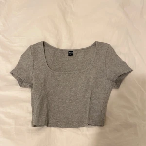 Grå croppad topp från SHEIN - En enkel och stilren grå croppad topp från SHEIN i storlek S med korta ärmar och rundad, djup urringning. Tillverkad i mjuk bomullsmix som känns skön mot huden. Perfekt att matcha med höga jeans eller kjol för en chill look.