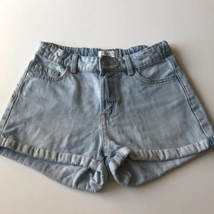 Ljusblå jeansshorts från Lindex - Snygga ljusblå jeansshorts från Lindex med klassisk femficksdesign och uppvikta benslut. Shortsen har bälteshällor, knapp och dragkedja framtill samt två bakfickor. Perfekta för varma dagar och enkel att matcha med allt.