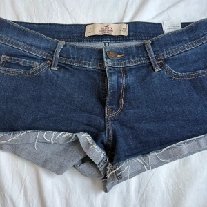 Mörkblå jeansshorts från Hollister - Snygga mörkblå jeansshorts från Hollister med råa kanter och klassiska fickor både fram och bak. Shortsen har normal passform, orange kontrastsömmar och är perfekta för sommaren. Materialet är jeans och de har låg midja för en avslappnad vibe.