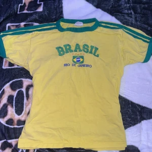 Gul Brasil t-shirt Rio de Janeiro - Gul t-shirt med gröna detaljer och texten 'BRASIL' samt 'RIO DE JANEIRO' broderat framtill. Klassisk sportig look med gröna ränder på axlarna och grönt runt hals och ärmslut. Perfekt för dig som älskar fotboll och Brasilien. (Köpt i rio de Janeiro)