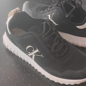 Skor - Snygga svarta sneakers från Calvin Klein med vit sula och stora CK-loggor på sidan. Ovandelen är i mesh och syntet, med svarta snören och vit detalj baktill där det står Calvin Klein. Perfekta för en stilren och sportig look.