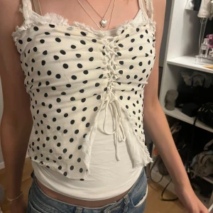 Prickig snörad linnetopp i vitt - Supersöt vit linnetopp med svarta prickar och snörning framtill. Toppen har råa kanter och croppad passform, perfekt att styla med jeans eller kjol. Lätt och luftig känsla, passar dig som gillar trendiga detaljer och lekfulla mönster.