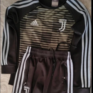 Juventus Adidas långärmad träningströja - Juventus långärmad träningströja från Adidas i svart med gråa ränder på ärmarna och grafiskt mönster framtill. Tröjan har rund hals, klassiska Adidas-loggan och Juventus-emblem på bröstet. Perfekt för fotbollsträning eller chill. Men byxornas dragkedja är sönder och byxorna har små hål men det är inget fel på tröjan 