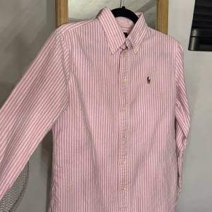 Rosa och vit randig skjorta Ralph Lauren - Säljer en snygg rosa och vit randig skjorta från Ralph Lauren, modell Custom Fit. Skjortan har klassisk krage, knäppning framtill och broderad logga på bröstet. Tillverkad i mjuk bomull och har långa ärmar. Perfekt för dig som gillar preppy stil!