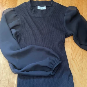 Svart ribbad tröja med puffärmar D-XEL - Supersnygg svart tröja från D-XEL med ribbad kropp och transparenta puffärmar i ett tunt material. Tröjan har rund halsringning och lång ärm med resår vid ärmslut. Perfekt för dig som vill ha en trendig och stilren look.