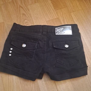 Svarta jeansshorts  - Svarta jeansshorts från WeTweens by KappAhl med snygga silverfärgade knappar och nitar på fickorna. Klassisk femficksmodell med uppvikta benslut och bälteshällor. Str 146