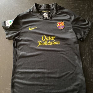 Svart FC Barcelona Nike fotbollströja - FC Barcelona bortatröja från Nike i svart med gula detaljer och klubbmärke på bröstet. Qatar Foundation och UNICEF-loggor tryckta i gult. Tillverkad i Dri-FIT-material som andas, perfekt för träning eller match. Klassisk krage och korta ärmar.