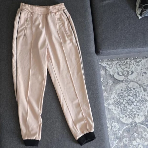 Beige mjukisbyxor med svarta detaljer - Snygga beige mjukisbyxor med svarta muddar och svarta ränder längs sidorna. Byxorna har resår i midjan, dragkedjefickor framtill och är tillverkade i ett mjukt material som känns skönt mot huden. Det finns en liten fläck på nedre delen av byxbenen (syns på bilden 4). Annars helt och fräsch 