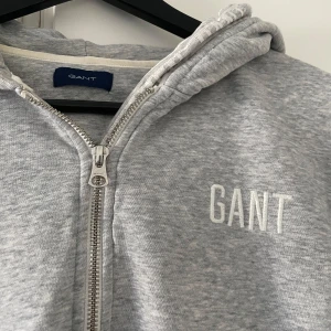 Grå hoodie från GANT med dragkedja - Snygg grå hoodie från GANT med halv dragkedja och broderad logga på bröstet. Tröjan har huva med tryckt GANT-text, känguruficka och långa ärmar. Perfekt för chill dagar eller när du vill ha en clean och sportig look.