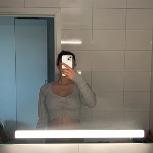 Grå croppad stickad tröja med snörning - Superfin ljusgrå croppad stickad tröja med v-ringning och långa ärmar. Tröjan har snygg snörning framtill som ger en cool rynkad effekt. Perfekt till jeans eller kjol för en trendig look.