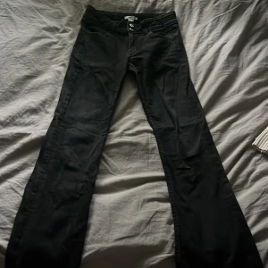 Svarta bootcut jeans från Gina Tricot - Snygga svarta jeans från Gina Tricot med bootcut passform och låg midja. Jeansen har dubbla knappar i midjan, klassiska fem fickor och diskreta sömdetaljer på bakfickorna. Perfekta för dig som gillar en stilren look med lite extra edge.