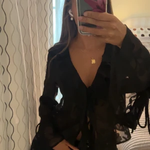 Svart transparent blus med volang och snörning - Supersnygg svart transparent blus från Gina med volangdetaljer och snörning framtill. Vida ärmar med knytband och lätt blommönstrad struktur i tyget. Perfekt för dig som vill sticka ut med en unik och trendig look.