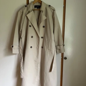 Beige trenchcoat från Vero Moda - Stilren beige trenchcoat från Vero Moda med klassisk dubbelknäppning och bälte i midjan. Jackan har långa ärmar med knappar vid ärmslut och två fickor i sidan. Perfekt för lager-på-lager och snygg till alla outfits. Sparsamt använd. Strl S (normal i storlek). Liten fläck på baksidan, se bild.