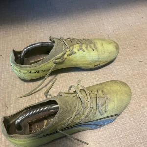 Gula Puma Ultra fotbollsskor - Säljer ett par snabba Puma Ultra fotbollsskor i gul färg med blå detaljer och svart logga. Skorna har snörning och ovandel i syntetmaterial för lätt känsla på planen. Sulan har fasta dobbar i blått och svart, perfekt för gräsplaner. Riktigt snygg och sportig modell! Dom är inte sönder bara smutsiga