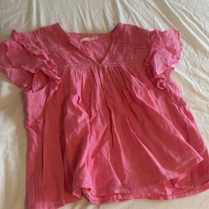 Rosa blus med volangärmar Gina Tricot - Supersöt rosa blus från Gina Tricot med v-ringning och volangiga korta ärmar. Blusen har smockdetaljer upptill och är luftig med en flowy passform. Perfekt för dig som gillar en feminin och avslappnad stil. Lappen där det står ginatricot är av på halva sidan annars är den hel 