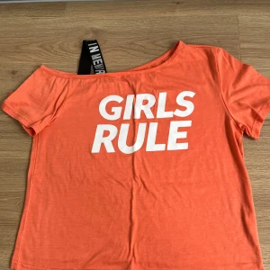 Orange offshoulder topp GIRLS RULE - Cool orange topp med offshoulder-design och korta ärmar. Framsidan har ett stort vitt tryck med texten 'GIRLS RULE'. En svart axelrem med vit text ger en extra detalj. Perfekt för dig som vill sticka ut med en färgstark och trendig look.