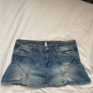 Blå jeanskjol med slitningar - Säljer en blå jeanskjol i kort modell med låg midja och coola slitningar framtill och baktill. Kjolen har klassiska fickor, bälteshällor och knappstängning. Perfekt för dig som gillar en avslappnad och trendig stil.
