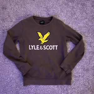 Brun sweatshirt från Lyle & Scott - Snygg brun sweatshirt från Lyle & Scott med stor gul logga och vit text på bröstet. Klassisk rund halsringning och långa ärmar. Perfekt för dig som gillar stilrena och sportiga plagg med tydlig branding.