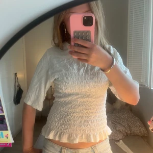 Vit smockad blus med volangkant - Supersöt vit blus med smockad midja och volangkant nertill. Toppen har halvlånga ärmar och en lätt rynkad struktur som ger en cool och trendig look. Perfekt att styla med jeans eller kjol för en fräsch vibe.