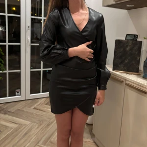 Svart omlottklänning i skinnlook - Snygg svart kort omlottklänning från Chiara Forthi i skinnimitation. Klänningen har långa puffärmar med knappdetalj vid ärmslut, markerad midja och v-ringning. Perfekt för dig som vill sticka ut med en edgy och trendig stil.