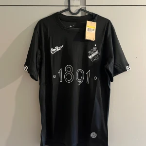 Svart AIK fotbollströja Nike 1891 - AIK fotbollströja i svart från Nike med tryckta detaljer som klubbmärke, årtalet 1891 och texten 'SMOKINGLIRARE' på ryggen. Kortärmad modell i lätt och ventilerande Dri-Fit-material. Snygga rutiga detaljer på ärmsluten.