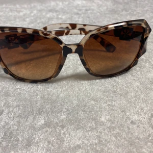 Dam Bruna mönstrade solglasögon  - Snygga solglasögon med brunmelerat mönster och stora, kantiga bågar. Glasen är bruna och bågarna har en cool silverdetalj vid sidorna. Perfekt accessoar för att lyfta din stil.