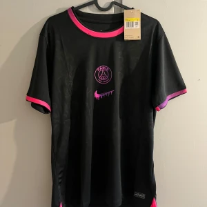 PSG x Nike svart fotbollströja - Svart PSG fotbollströja från Nike med rosa detaljer på krage och ärmslut. Paris Saint-Germain-logga och Nike swoosh i rosa framtill. Lätt och ventilerande material, perfekt för träning eller match. Snyggt mönster ton-i-ton i tyget och GTO-tryck i nacken.