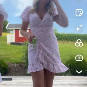 Ljusrosa omlottklänning med volang - Supersöt ljusrosa omlottklänning med små blommor, puffiga korta ärmar och volangkant nertill. Klänningen har v-ringning och knyts i midjan för en feminin siluett. Perfekt för sommarens alla tillfällen!