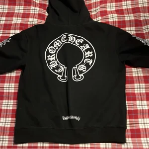 Svart Chrome Hearts hoodie med tryck - Svart hoodie från Chrome Hearts med stor vit logga på ryggen och mindre tryck på bröstet. Dragkedja framtill, snörning i huvan och detaljerade tryck längs ärmarna. Perfekt för dig som gillar streetwear och vill sticka ut.