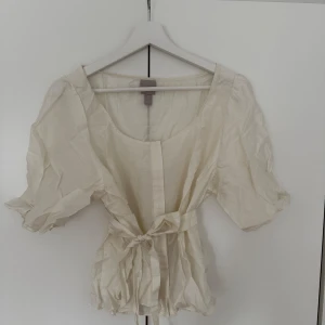 Krämvit blus med knyt i midjan - Supersöt krämvit blus från H&M med korta puffärmar och knytband i midjan. Blusen har en lätt glansig yta och en luftig känsla, perfekt för varmare dagar. Den har rund halsringning och en feminin, flowy passform.
