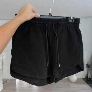 Svarta mjukisshorts - Svarta mjukisshorts från H&M med elastisk midja och snörning framtill. Loose fit med rundade benslut och tillverkad i en mix av 60% bomull och 40% polyester. Perfekta för chill dagar eller träning. Snygg och enkel design som funkar till allt. Önskar du veta exakta mått? Tveka inte att höra av dig!