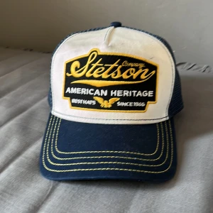 Stetson truckerkeps blå och vit - Snygg truckerkeps från Stetson med broderad gul och vit logga framtill. Kepsen är blå med vit frontpanel, gul kontrastsöm och justerbar snapback. Baksidan är i mesh för extra ventilation. Perfekt för dig som gillar klassisk amerikansk stil.