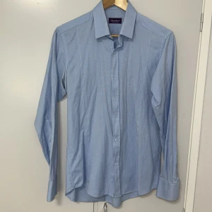 Ljusblå skjorta från Bernhard Exclusive - Klassisk ljusblå skjorta från Bernhard Exclusive med lång ärm och knappar framtill. Skjortan har en stilren krage och är tillverkad i ett mjukt bomullstyg som känns bekvämt mot huden. Perfekt för dig som gillar en clean och tidlös look.