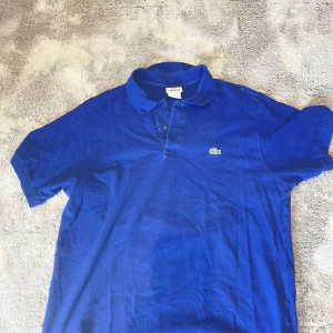 Blå pikétröja från Lacoste - Klassisk blå pikétröja från Lacoste med den ikoniska krokodilloggan på bröstet. Tröjan har korta ärmar, krage och knäppning framtill. Tillverkad i mjuk bomull som känns skön mot huden. Perfekt för en clean och somrig look!