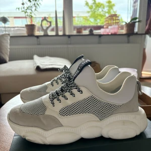 Moschino vita chunky sneakers - Säljer ett par vita chunky sneakers från Moschino med grå mockadetaljer, meshpaneler och svartvita snören. Sulan har en unik nallebjörnsdesign undertill och skorna har tydlig Moschino-logga på plösen. Perfekta för dig som vill sticka ut med en cool streetstyle. Nypris ungefär 3000kr, modellen säljs inte längre!