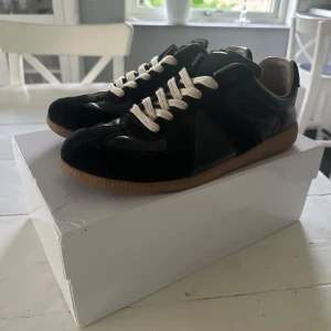 Maison Margiela svarta  - Maison Margiela sneakers i svart skinn och mocka med vita snören och beige gummisula. Klassisk low-top silhuett med rund tå och snygga paneler i olika material. Märkets logga syns på plösen och sulan. Perfekta för dig som gillar stilrena och lyxiga sneakers. Äktebevis finns tyvärr inte.
