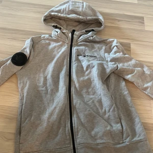 Grå hoodie från Sail Racing - Snygg ljusgrå hoodie från Sail Racing med svart logga på bröstet och stor text på ryggen. Tröjan har huva med snörning, hel dragkedja och ett svart märke på ena ärmen. Perfekt för dig som gillar sportig och avslappnad stil.