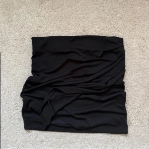 Svart tight kortkjol från Soda - Svart tight kortkjol från Soda med enkel och stilren design. Kjolen är stretchig och har en lätt draperad effekt på ena sidan. Perfekt för dig som gillar minimalistisk stil och vill ha en kjol som sitter snyggt på kroppen.
