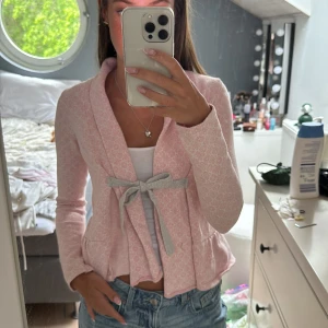 Odd Molly kofta - Supersöt ljusrosa cardigan jacka från Odd Molly med diskret mönster, grå knytrosett framtill och broderad logga på ryggen. Jackan har lång ärm, volangkant nertill och är gjord i mjuk bomullsmix. Perfekt för dig som gillar en feminin och chill stil.